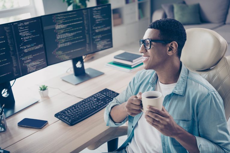 A man sitting in front off two monitors with lines of code, smiling